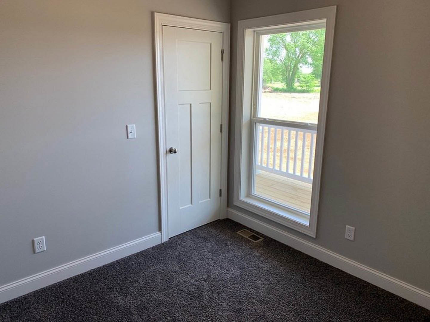 White door with silver handle, large window with white railing overlooking green trees, grey carpeted floor, white wall outlet on painted wall