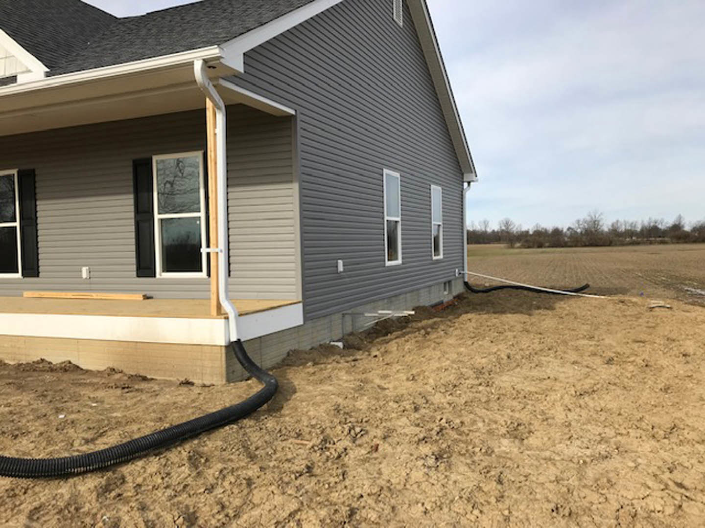 Tan siding house with white-framed window, black hose along ground, metal gutter, unfinished dirt yard, metal structure in foreground, gray shingle roof under blue sky