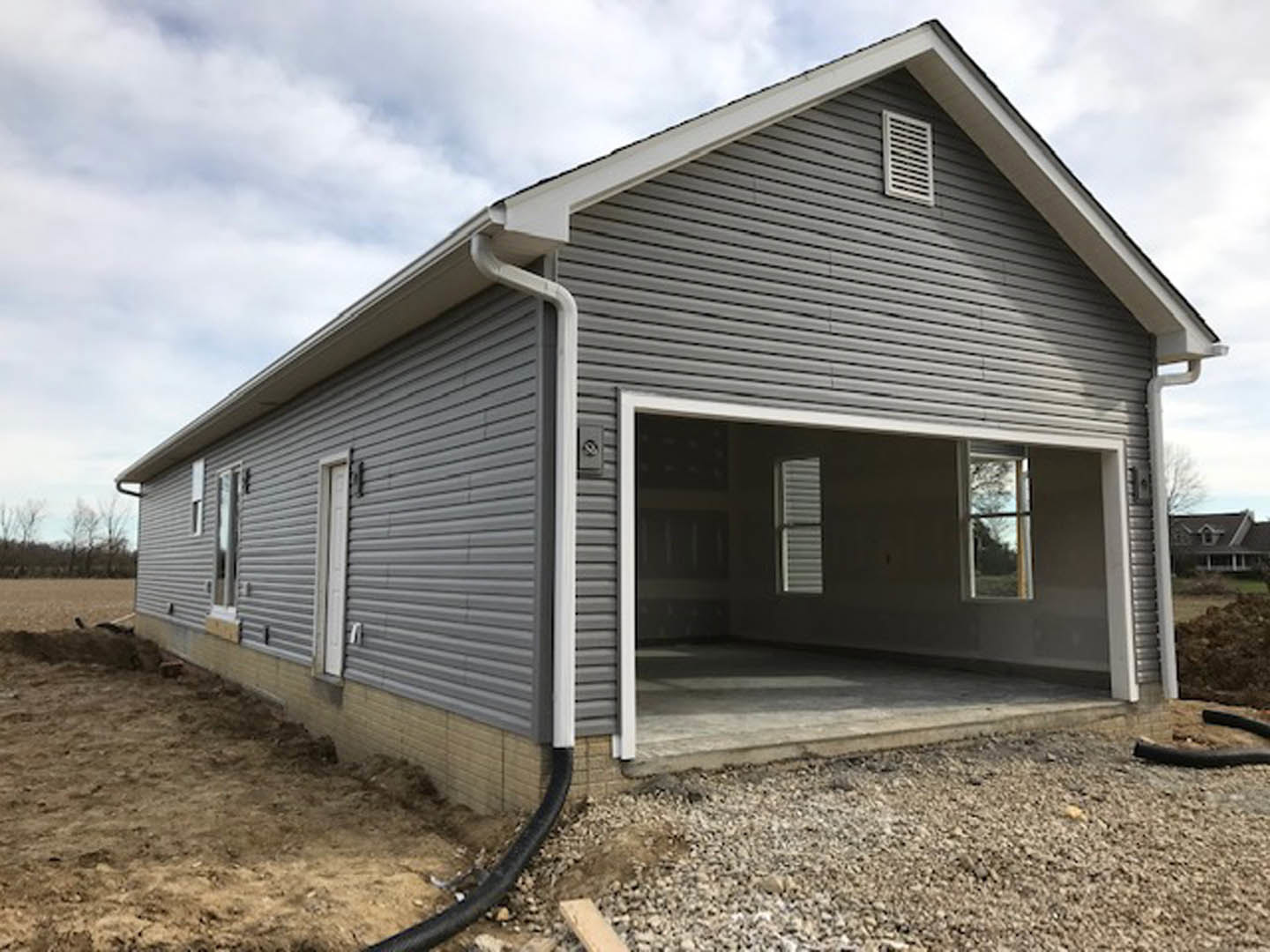 Grey siding house under construction with attached garage, white vent, windows with blinds, cloudy sky overhead
