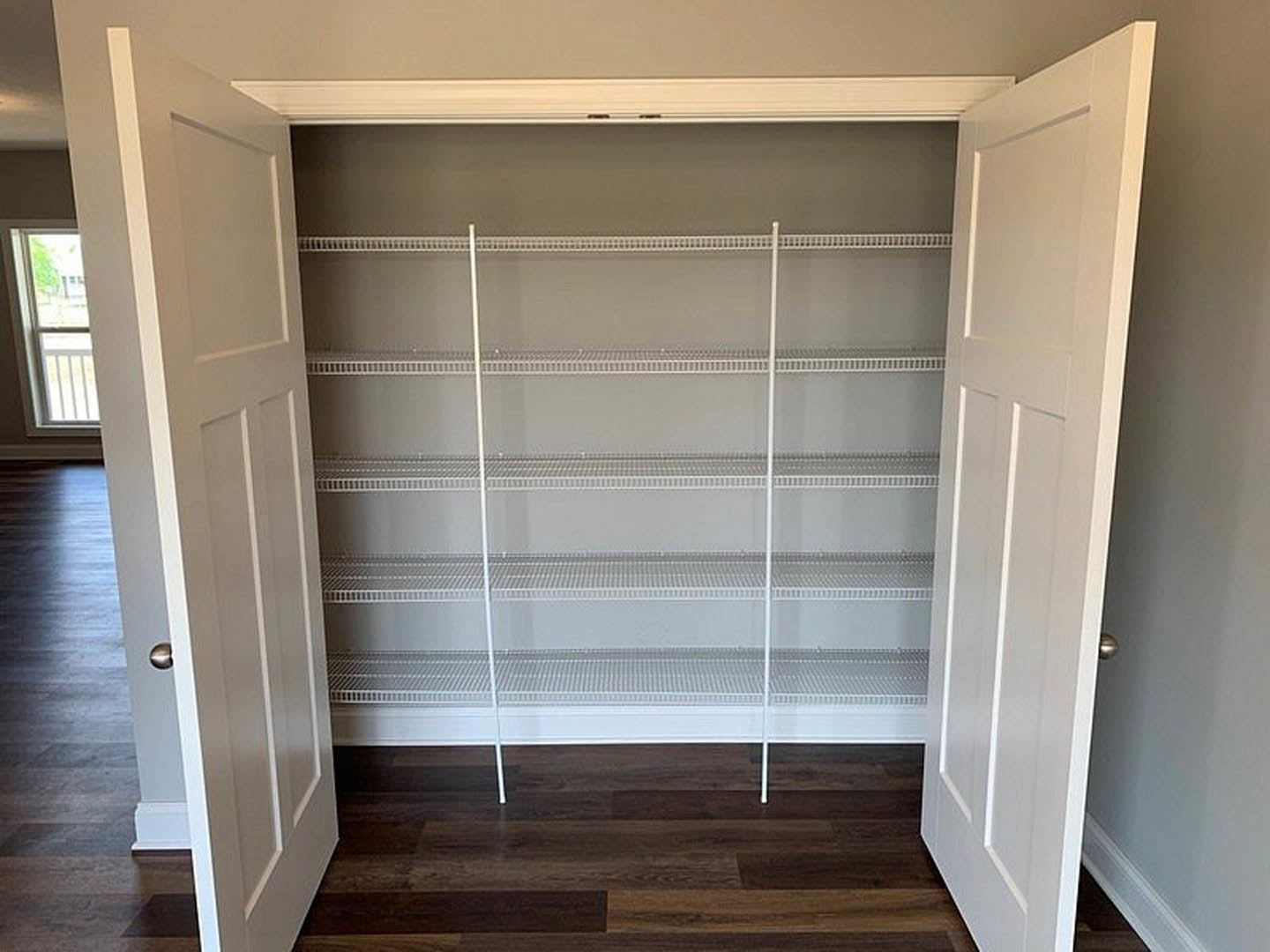 Walk-in closet with open white doors, built-in white shelving, hardwood floor, and a window with a white frame