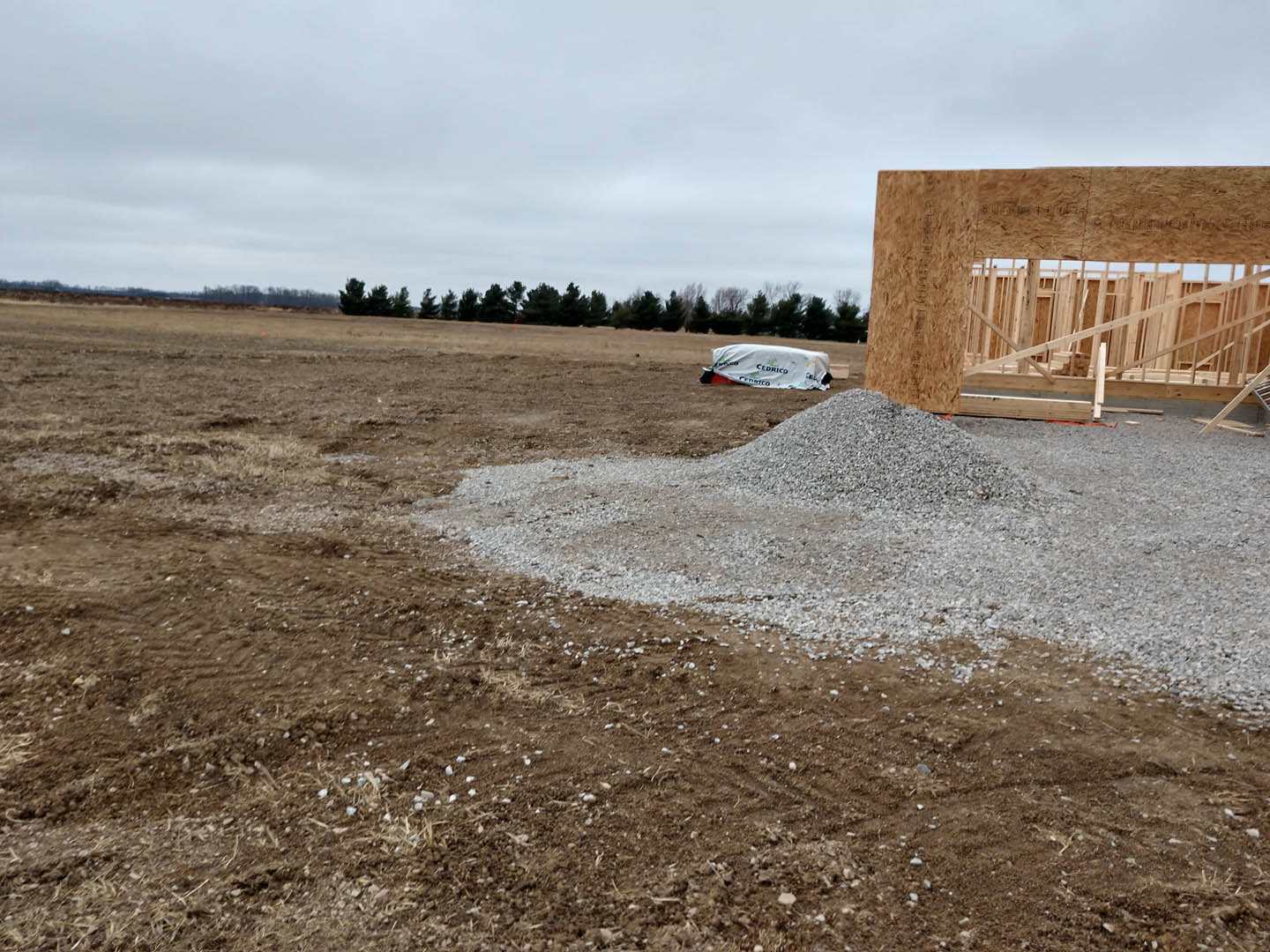 Wood-framed house under construction with white weather barrier, pile of gravel beside support pole, cloudy sky overhead, bare soil and grass surrounding site