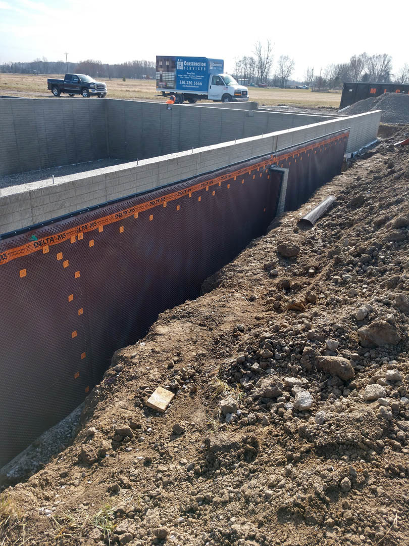 Excavated construction site with exposed soil, large pit, brick wall, parked trucks, and grassy surroundings under open sky