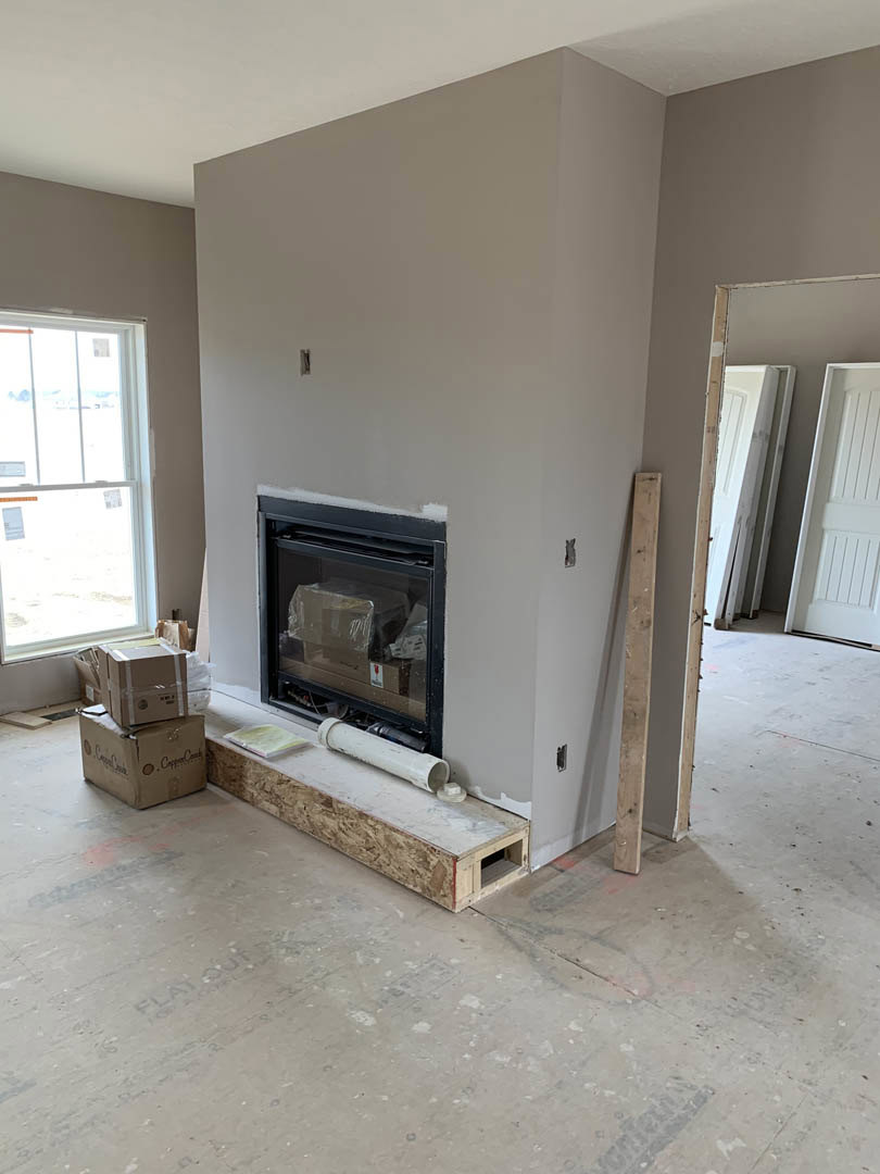 Stone fireplace with glass door, white-framed window, white paneled door, wood flooring, and white pipe on wooden surface in a plaster-walled living room