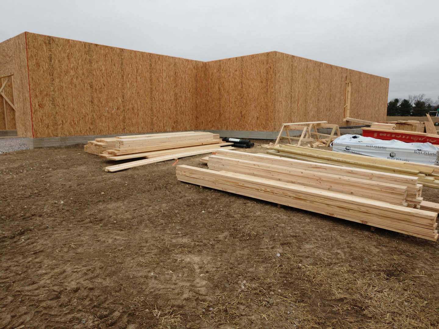 Partially built house with exposed wood framing, stacks of lumber on dirt ground, white construction bag with black text, clear sky in background