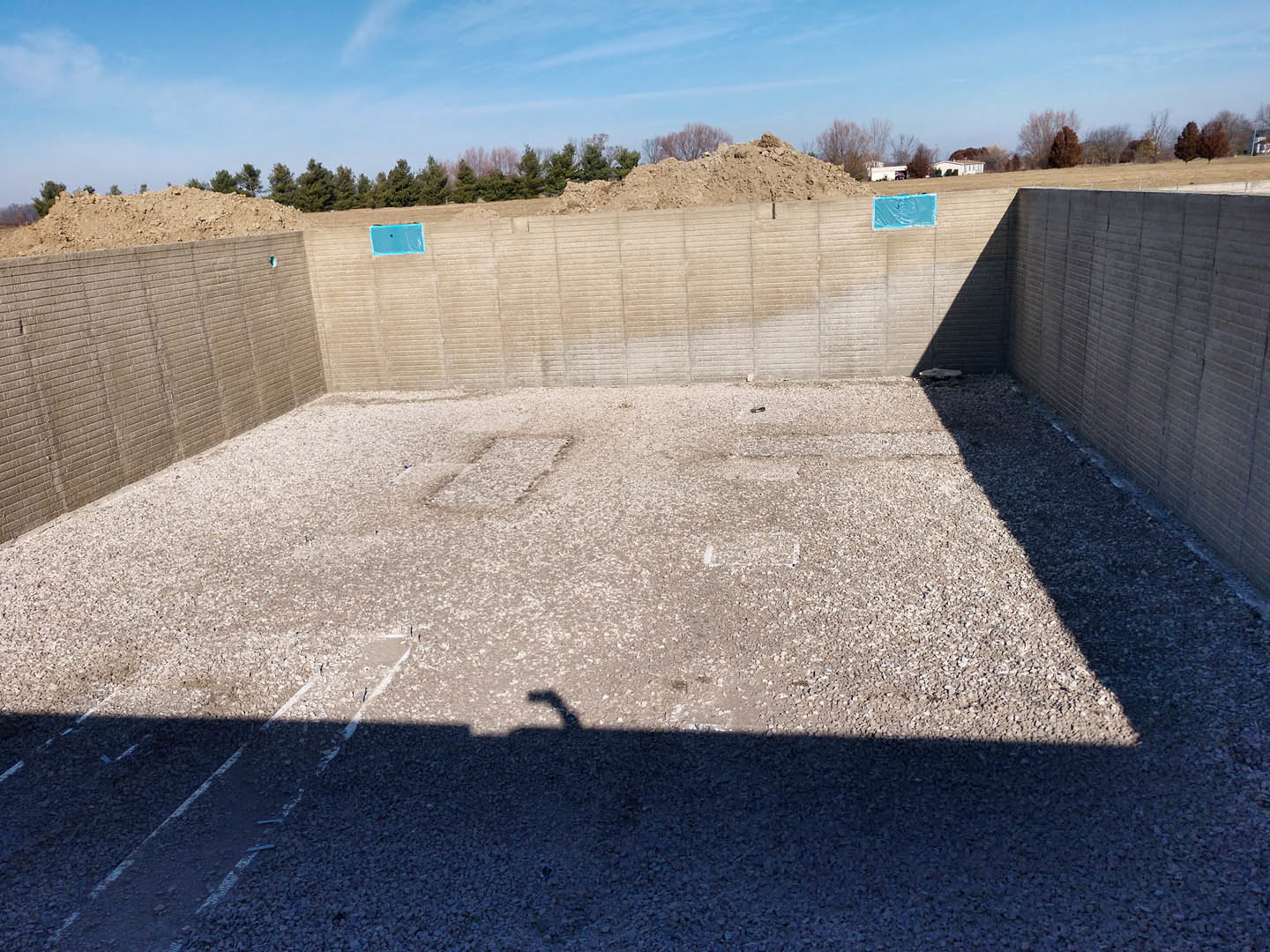 Concrete foundation surrounded by sandy soil and a pile of dirt, blue tarp partially covering ground, trees and blue sky with wispy clouds in background, shadow of a hand cast on