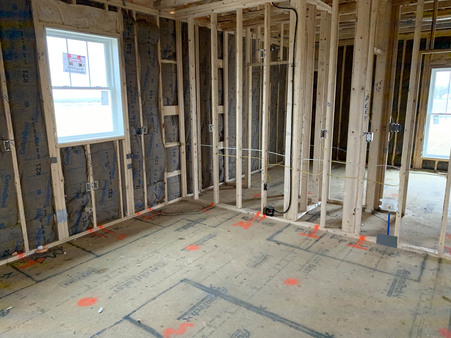 Wood-framed room under construction with exposed beams, window featuring a posted sign, and orange markings on subfloor
