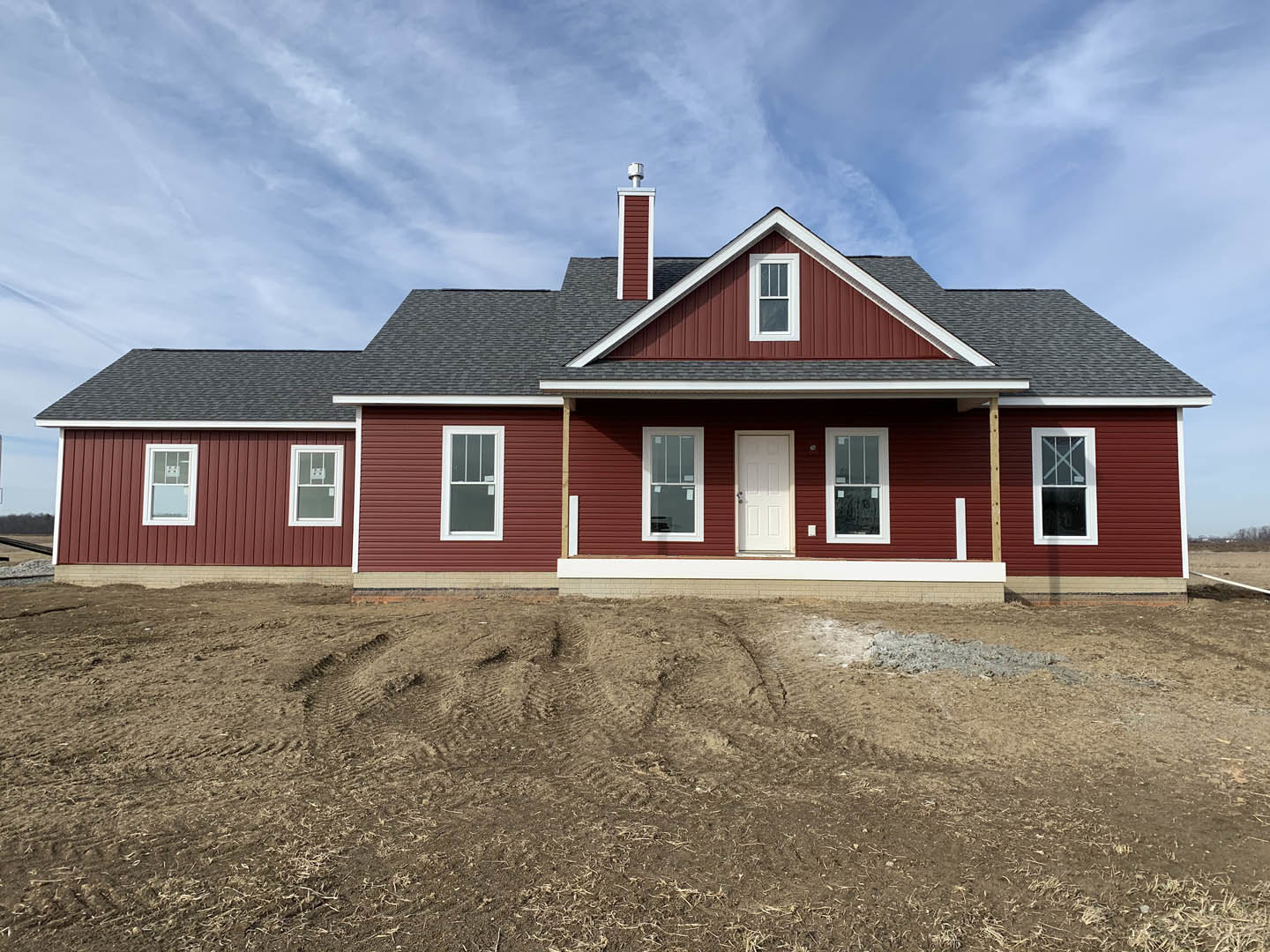Single-story house with white door and white-framed windows, brick chimney, dirt yard in foreground, red building visible in background, partly cloudy sky overhead