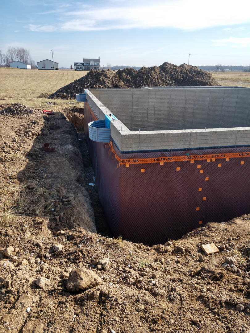 Excavated foundation pit surrounded by soil and construction materials, with partially built concrete walls and a blue container nearby; residential house and tower visible in the