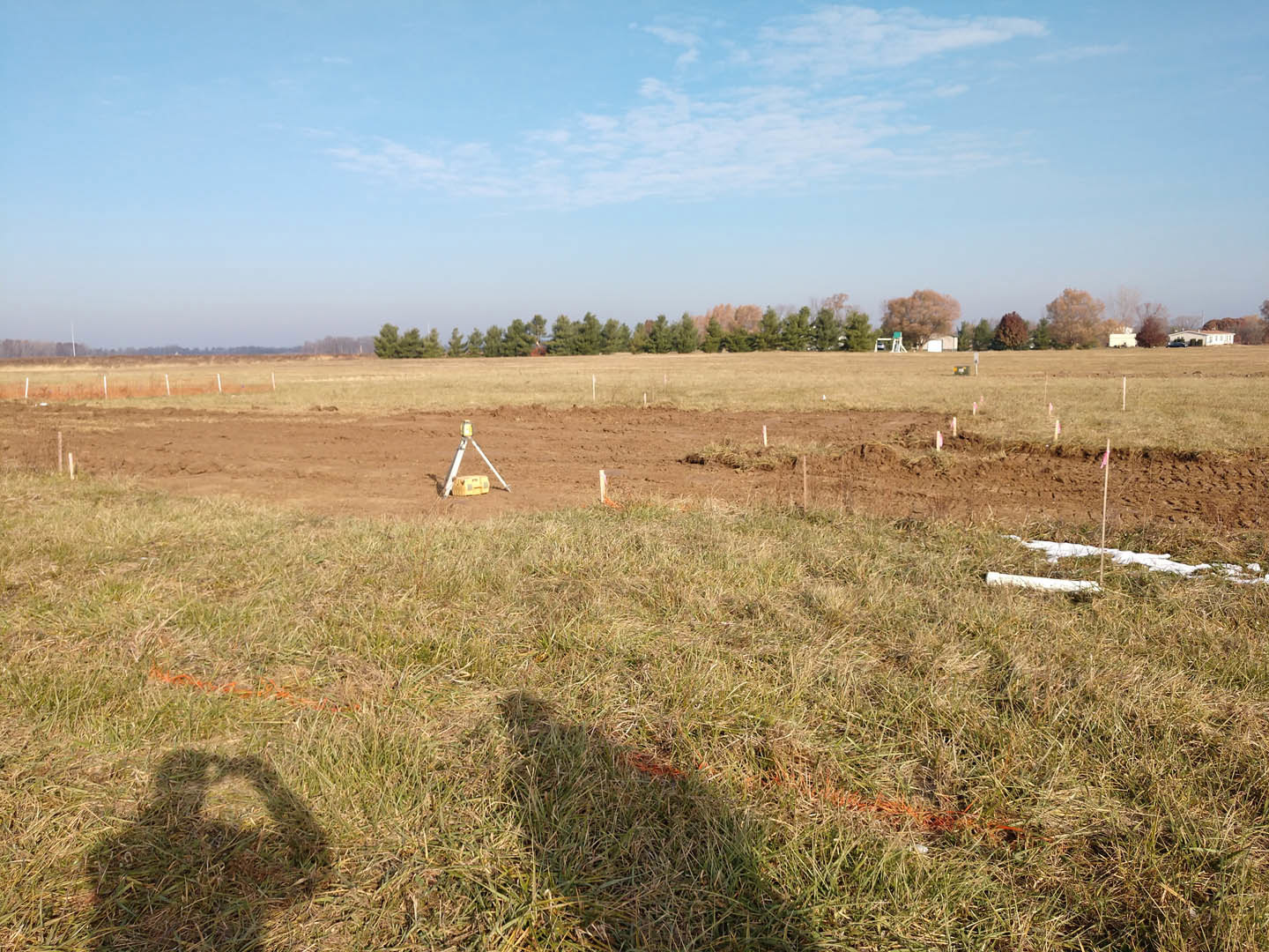 Dirt field bordered by trees with brown leaves, grassy patches, blue sky with scattered clouds, yellow tripod object, and shadow of a person on the grass