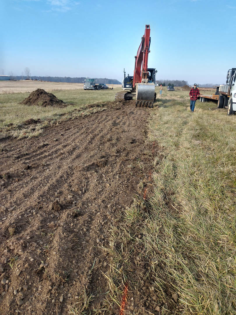 Red tractor parked on grassy field under clear blue sky, soil and dirt piles nearby, man standing at rear of machine.