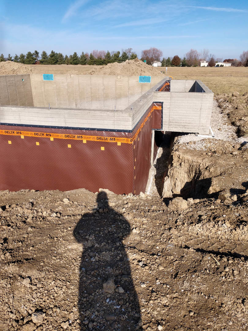 Shadow of a person cast on dirt field near a building under construction with red wall, leafless tree, and blue sky with scattered clouds