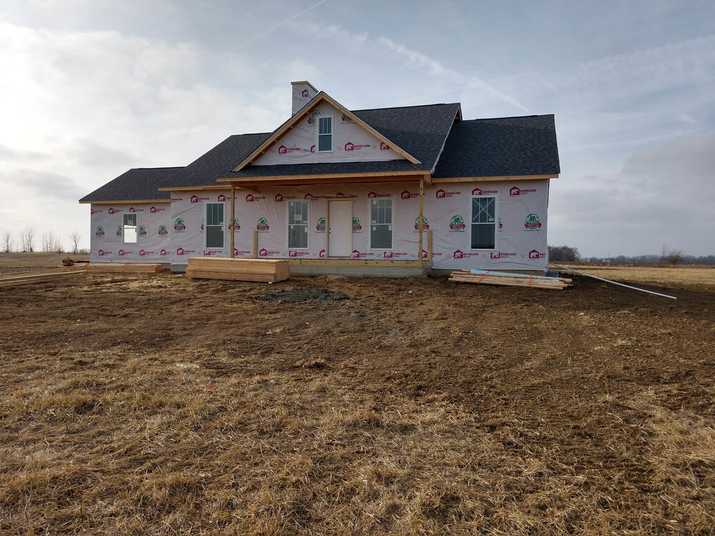 Partially built farmhouse with white framed windows, plastic sheeting covering exterior walls, grassy field in foreground, pile of lumber near entrance, white door with black knobs
