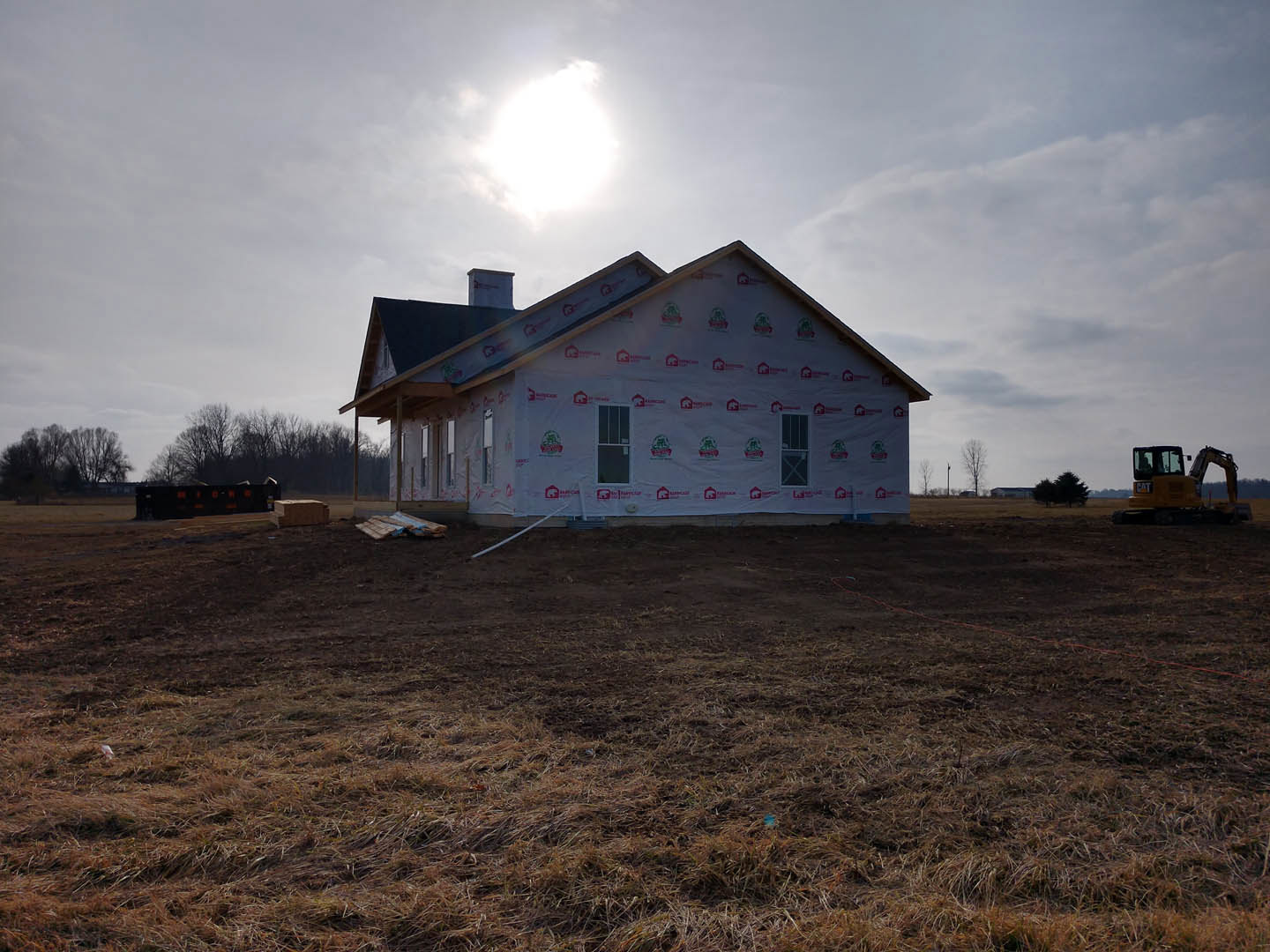 Two-story house with white-framed windows, covered roof, and dirt field in foreground; distant building and trees under cloudy sky.