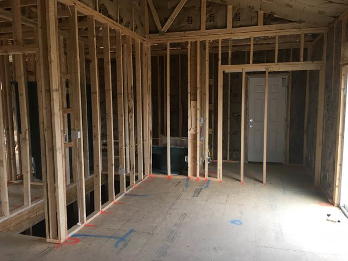 Unfinished room with exposed wood framing, visible ceiling beams, and construction materials; white door installed in framed wall.