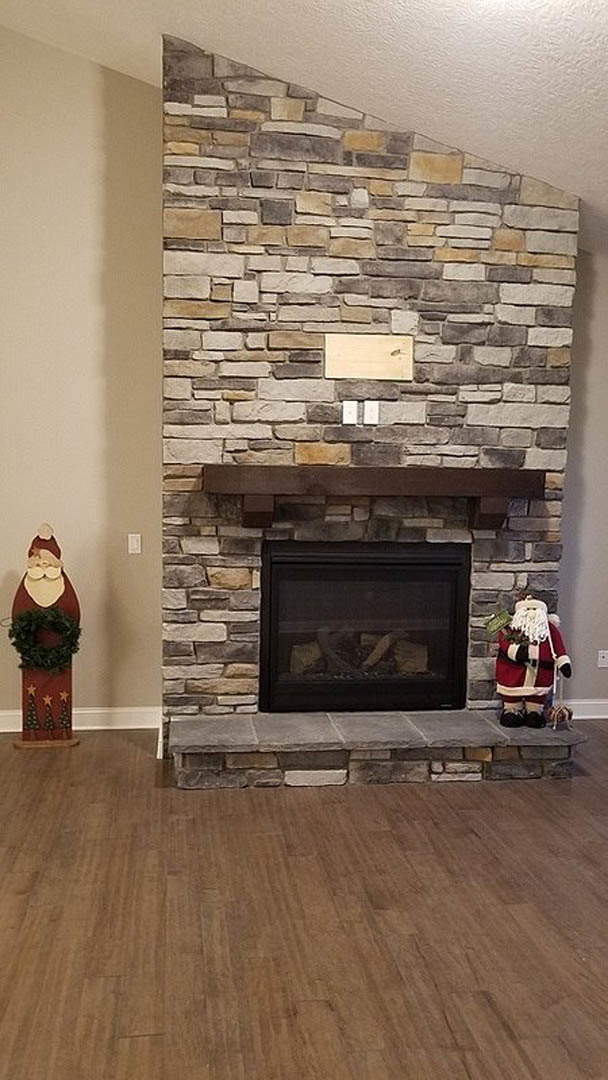 Stone fireplace with glass door and wood mantle, burning fire inside, Christmas wreath hanging above, hardwood floor with stone steps, man in Santa suit standing nearby
