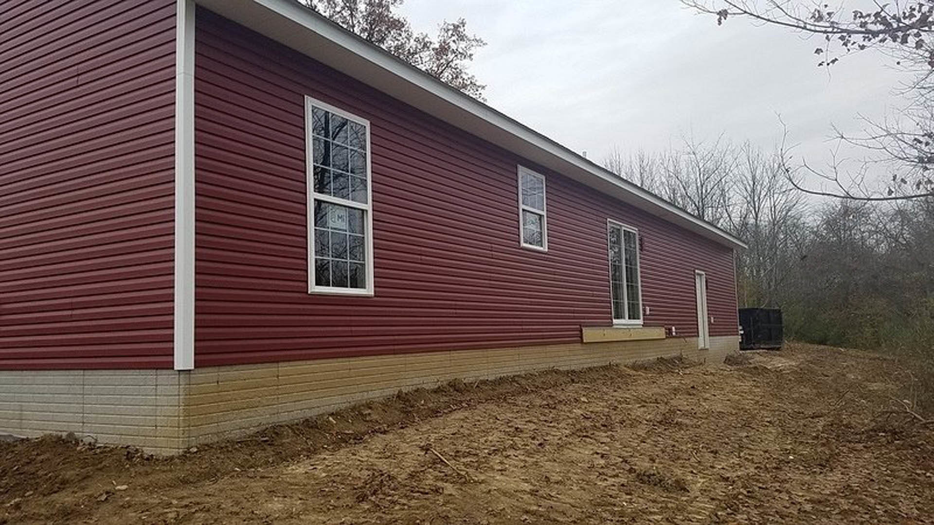 Red house with white trim, dirt patch in front yard, white post attached to red siding, window displaying a sign, tree and sky in background.