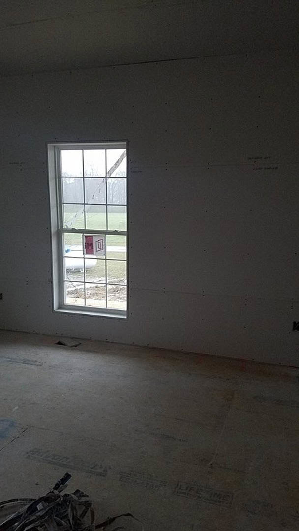 Large window set in a plaster wall, daylight streaming into an empty room with scattered black and white objects on the floor, minimal fixtures visible.