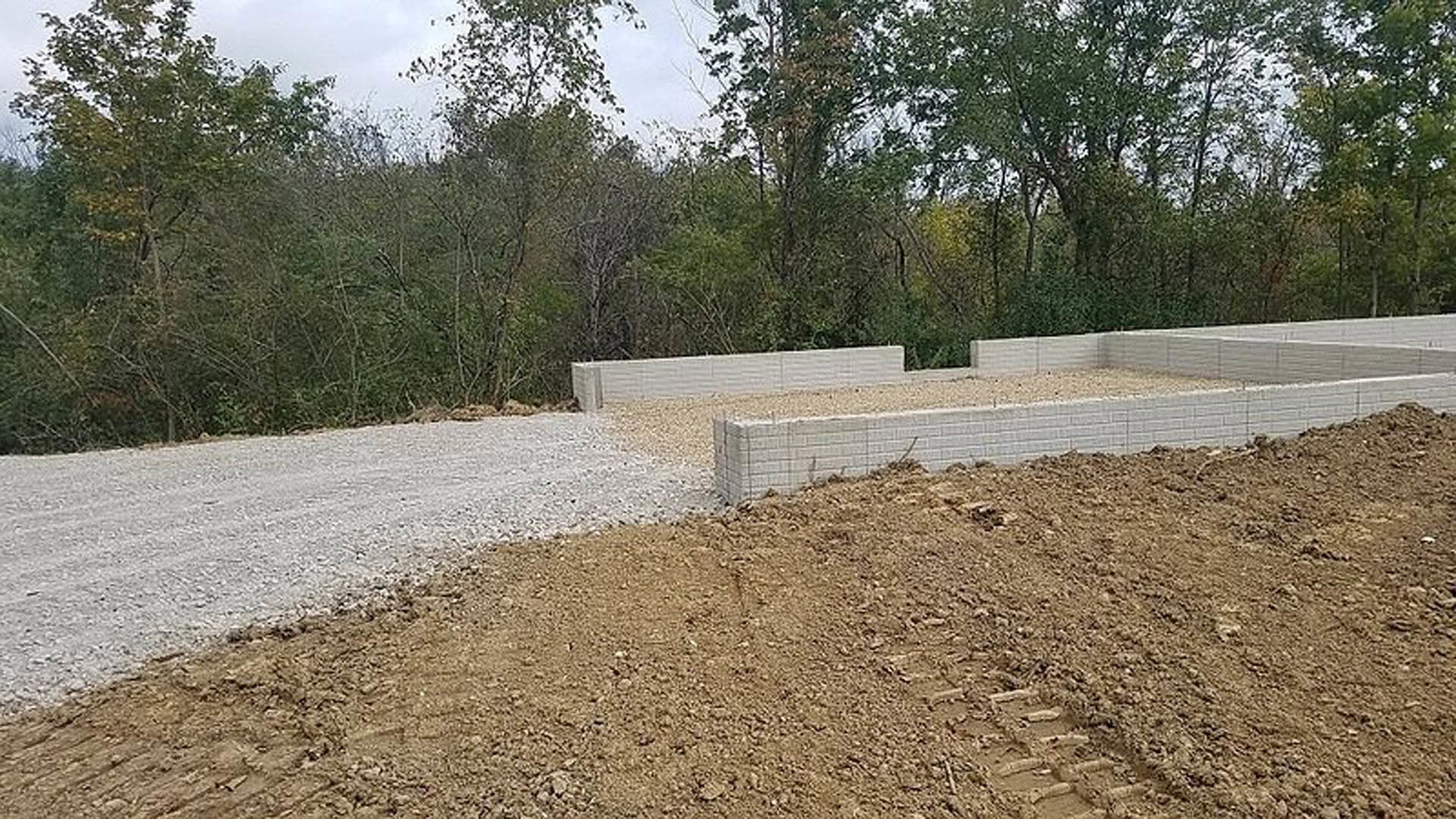 Dirt road with tire tracks leading past a partially built brick wall, surrounded by trees and construction materials on a land lot