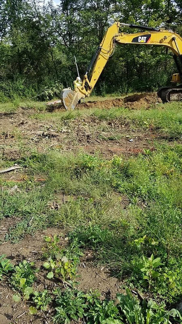 Yellow excavator digging a hole in grassy field with trees in background and exposed soil