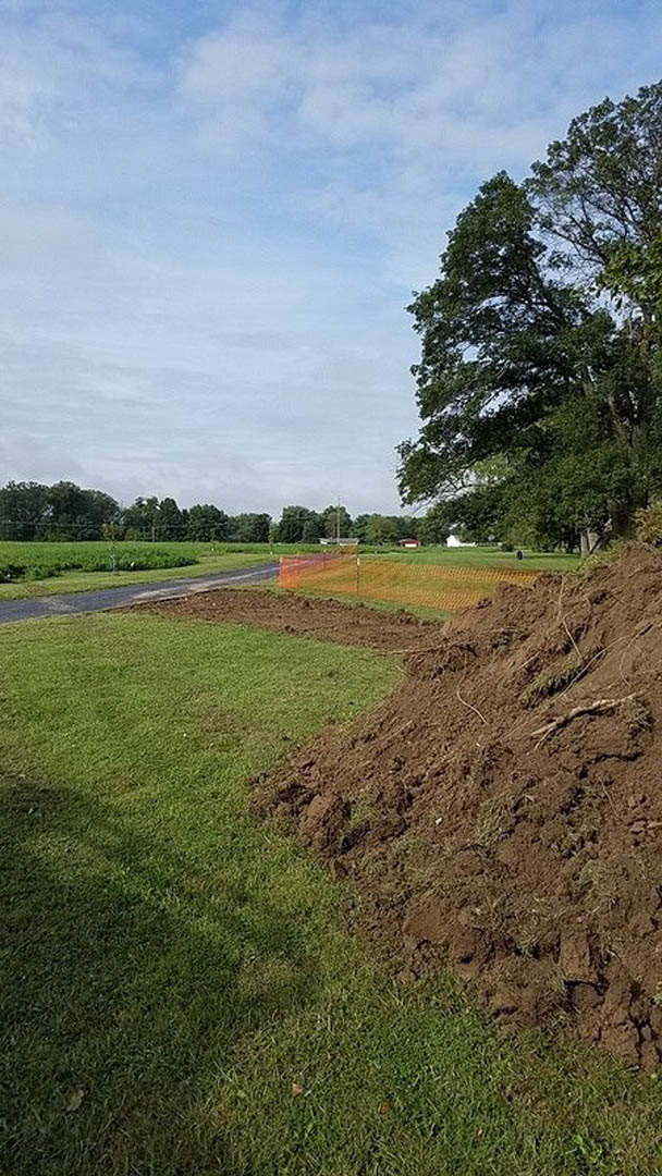 Dirt mound surrounded by grassy field, leafy tree, wooden fence, and cloudy sky in rural landscape