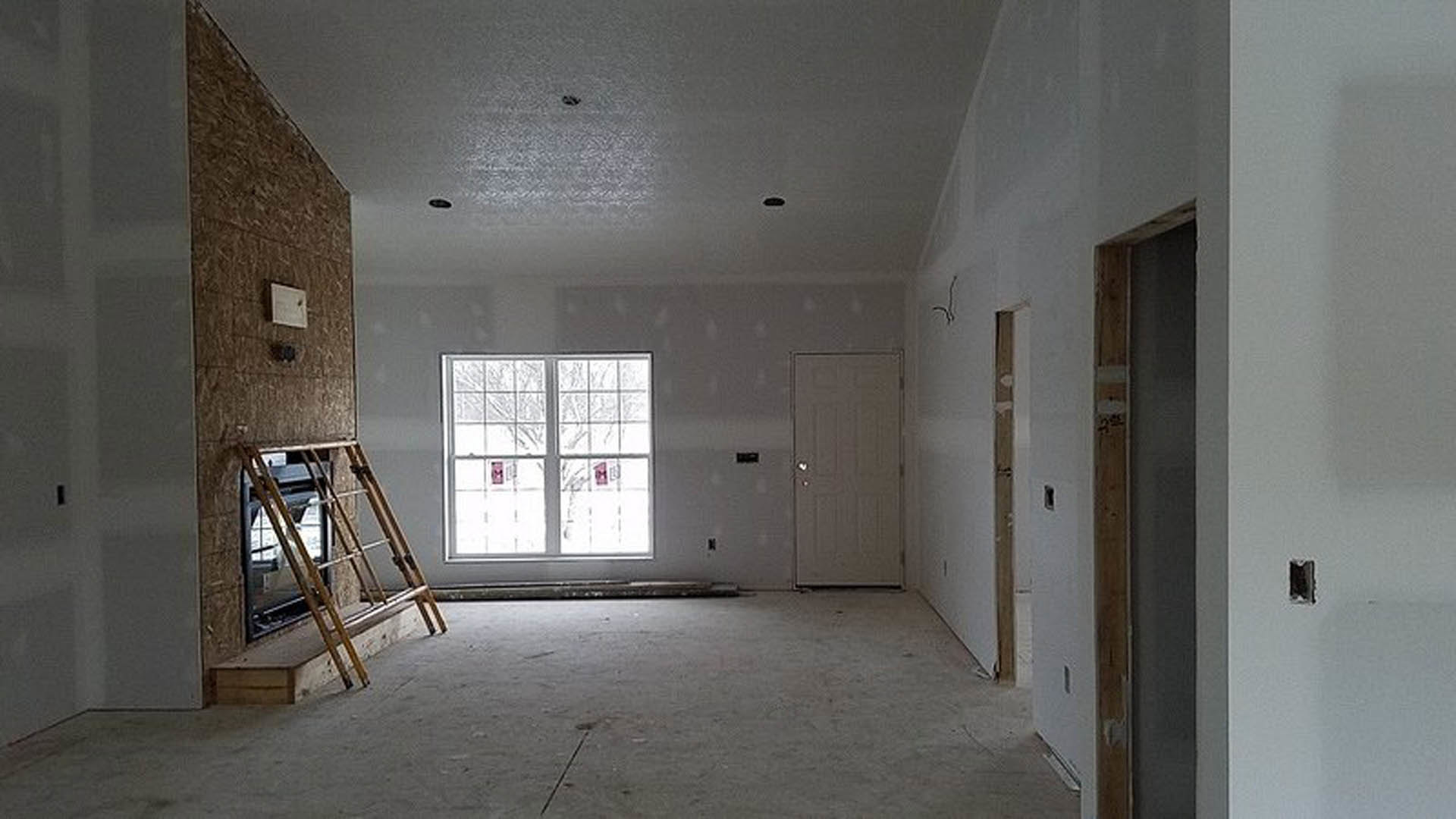 Living room with plaster walls, wood flooring, fireplace, ladder leaning against hearth, large window with signage, white thermostat, and black vent on wall.