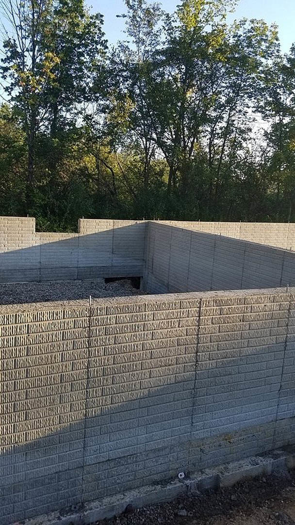 Concrete exterior wall with circular opening in the ground, wire fencing and trees visible in background, sunlight casting shadows on textured surface.