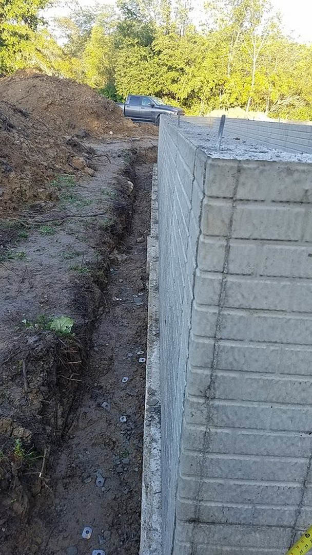 Concrete perimeter wall topped with metal fence, bordered by trees and plants, adjacent to paved driveway