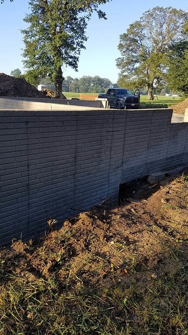 Black pickup truck parked beside red brick wall, grassy lawn and mature tree in background, clear blue sky overhead