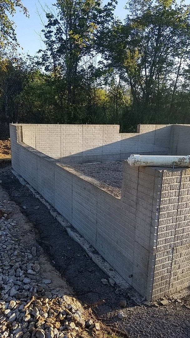 Concrete exterior wall with exposed pipe, gravel foundation, and scattered rocks on the ground; white brick wall partially visible with shadow.