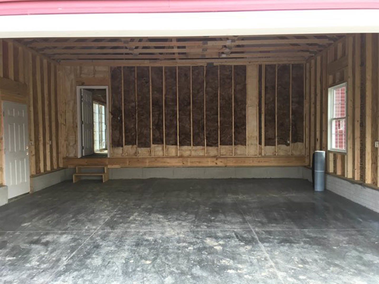 Concrete floor with close-up of wood panel door, grey metal pole with white stripe, wall featuring wooden window with bars, and ceiling beams.