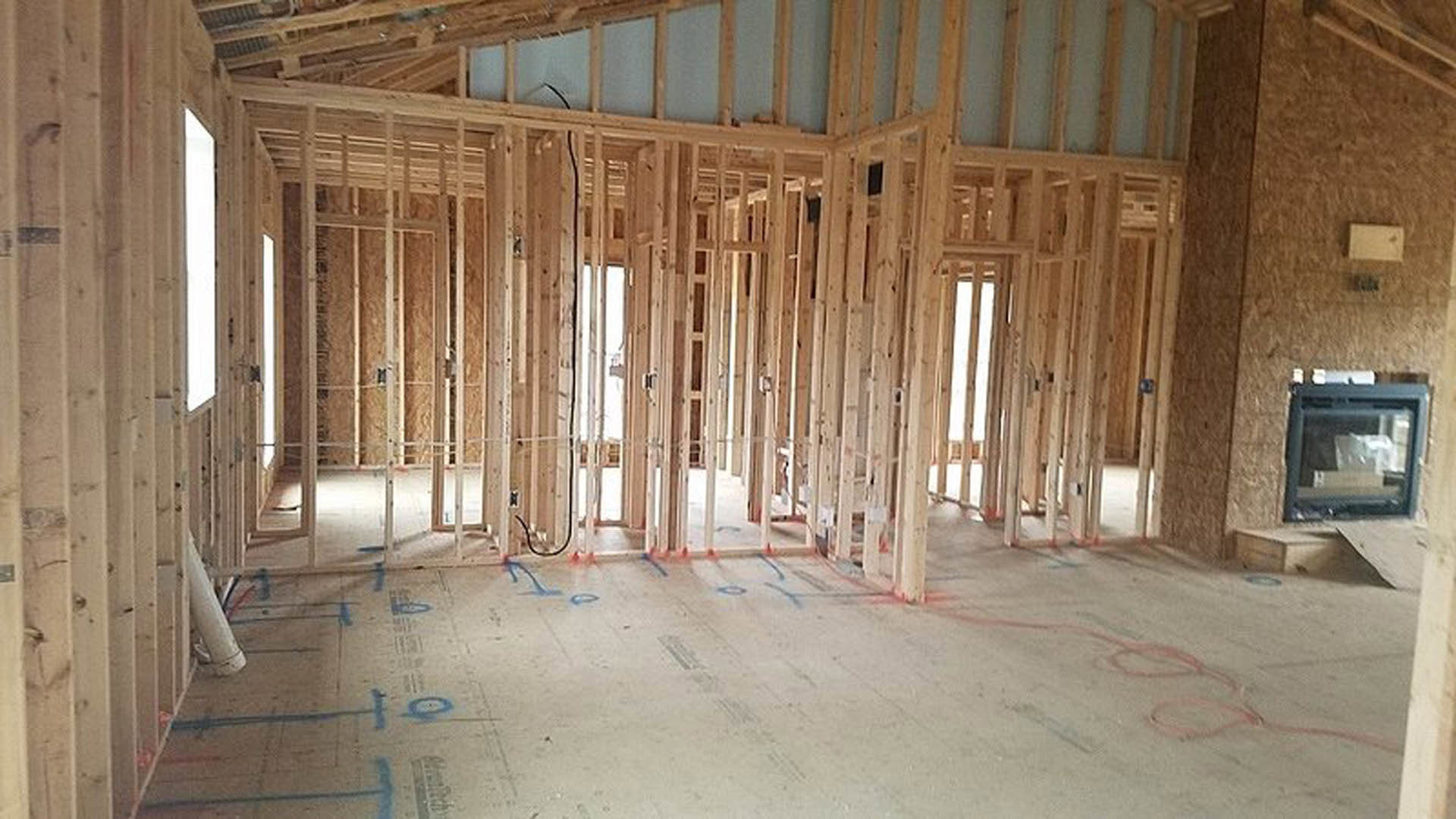 Wood-framed room under construction with exposed beams, concrete floor marked with blue paint, red hoses, and architectural blueprints laid out