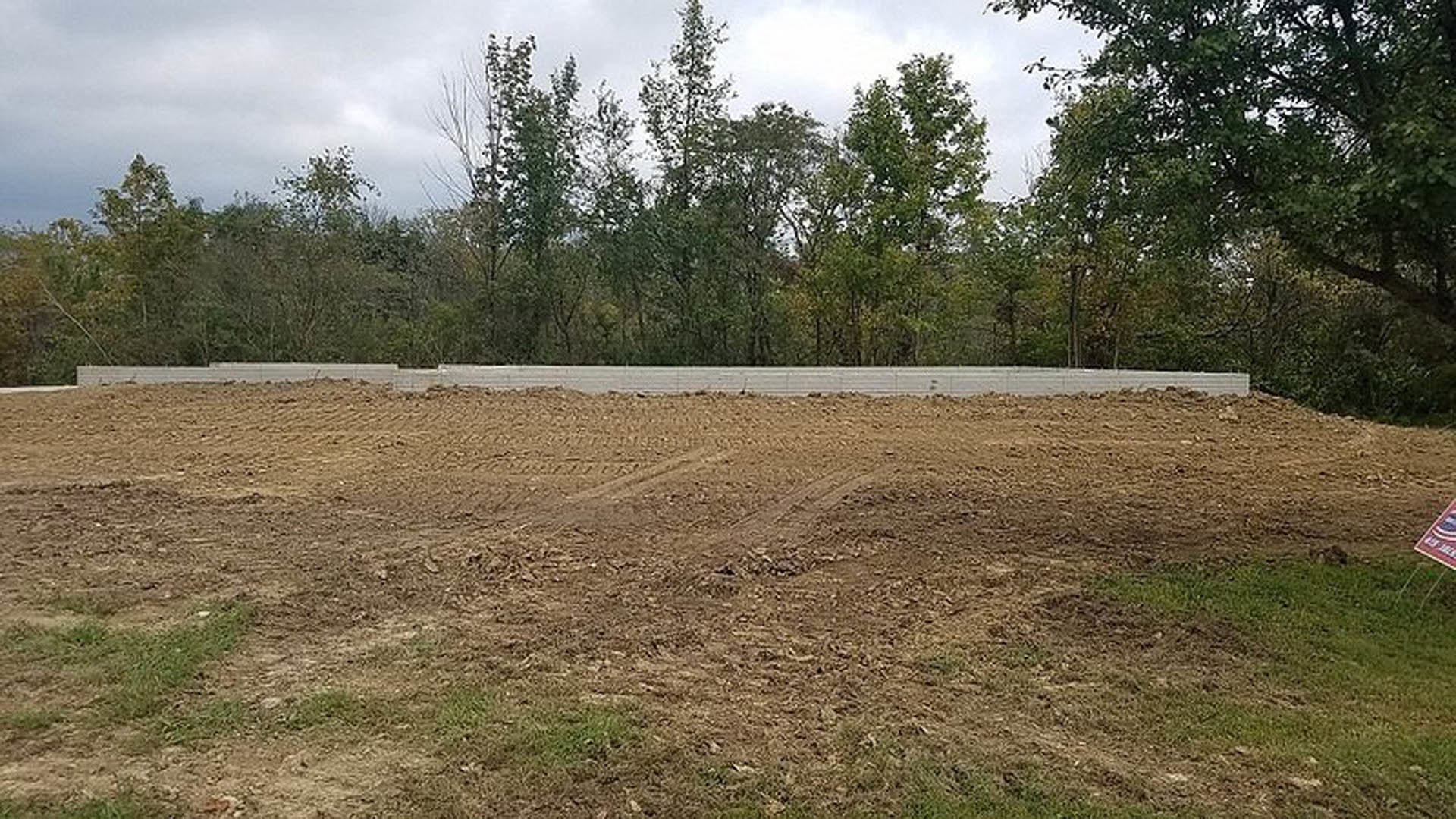 Dirt field bordered by a white fence, tire tracks visible in soil, group of leafy trees in background under cloudy sky