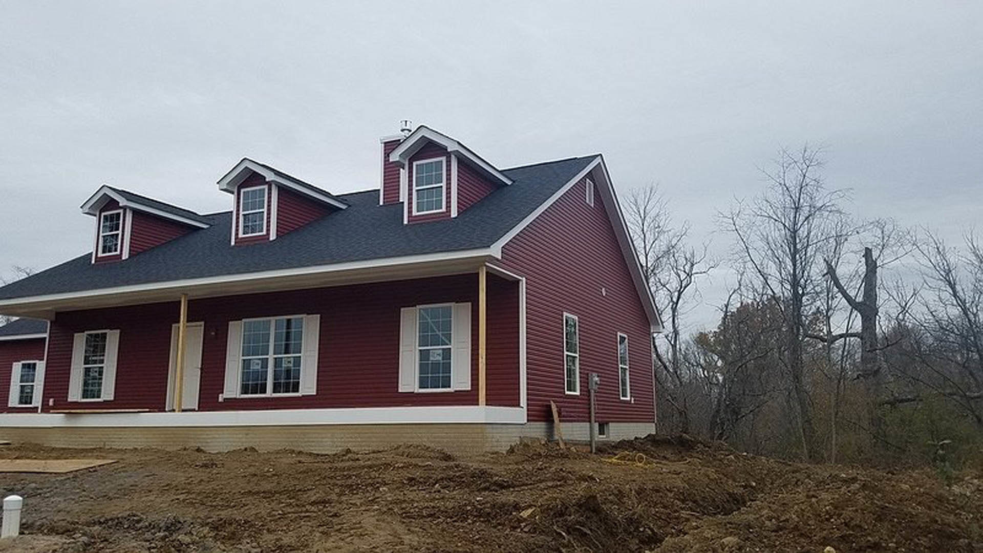 Partially built house with red metal roof, white-framed windows with multiple panes, temporary sign taped to glass, surrounded by tall trees and natural woodland setting