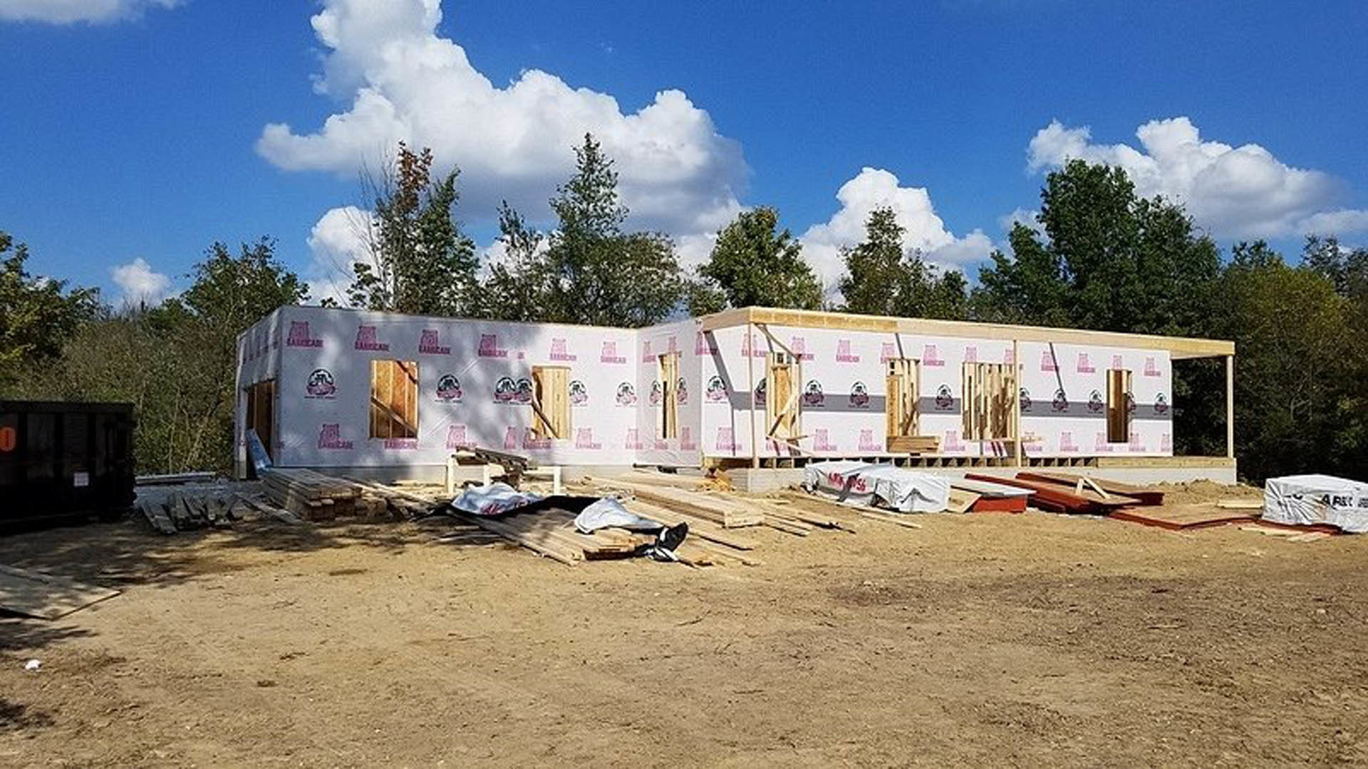 Framed residential structure under construction with exposed wood planks, surrounded by trees and cloudy sky