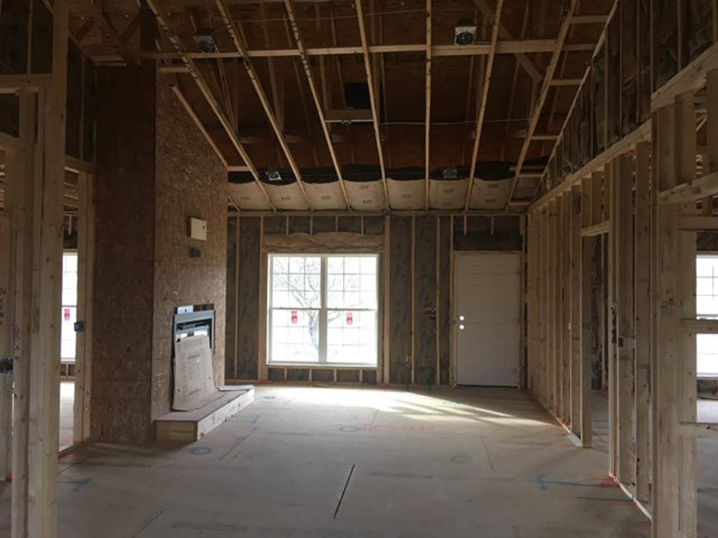 Living room with exposed wooden ceiling beams, stone fireplace, large window with tree view, and white door with sidelights.