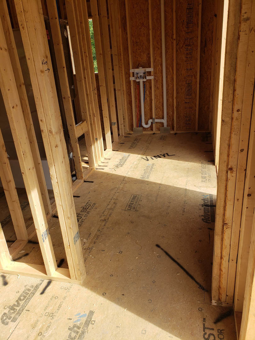 Unfinished room with exposed wood framing, vertical pipe installed in wall, plywood subfloor, and construction materials visible