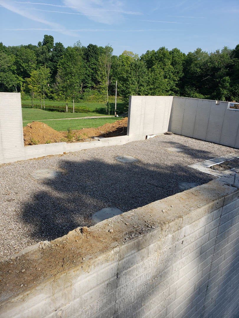 Concrete foundation and slab with gravel, partially built white wall featuring a drain, surrounded by trees under a blue sky with scattered clouds