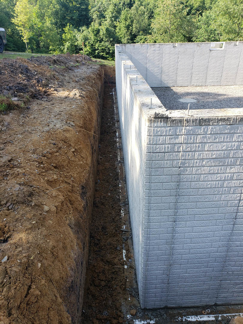 Concrete wall with deep trench in gravel foreground, white brick wall partially shaded, trees in background, manhole cover visible on ground.