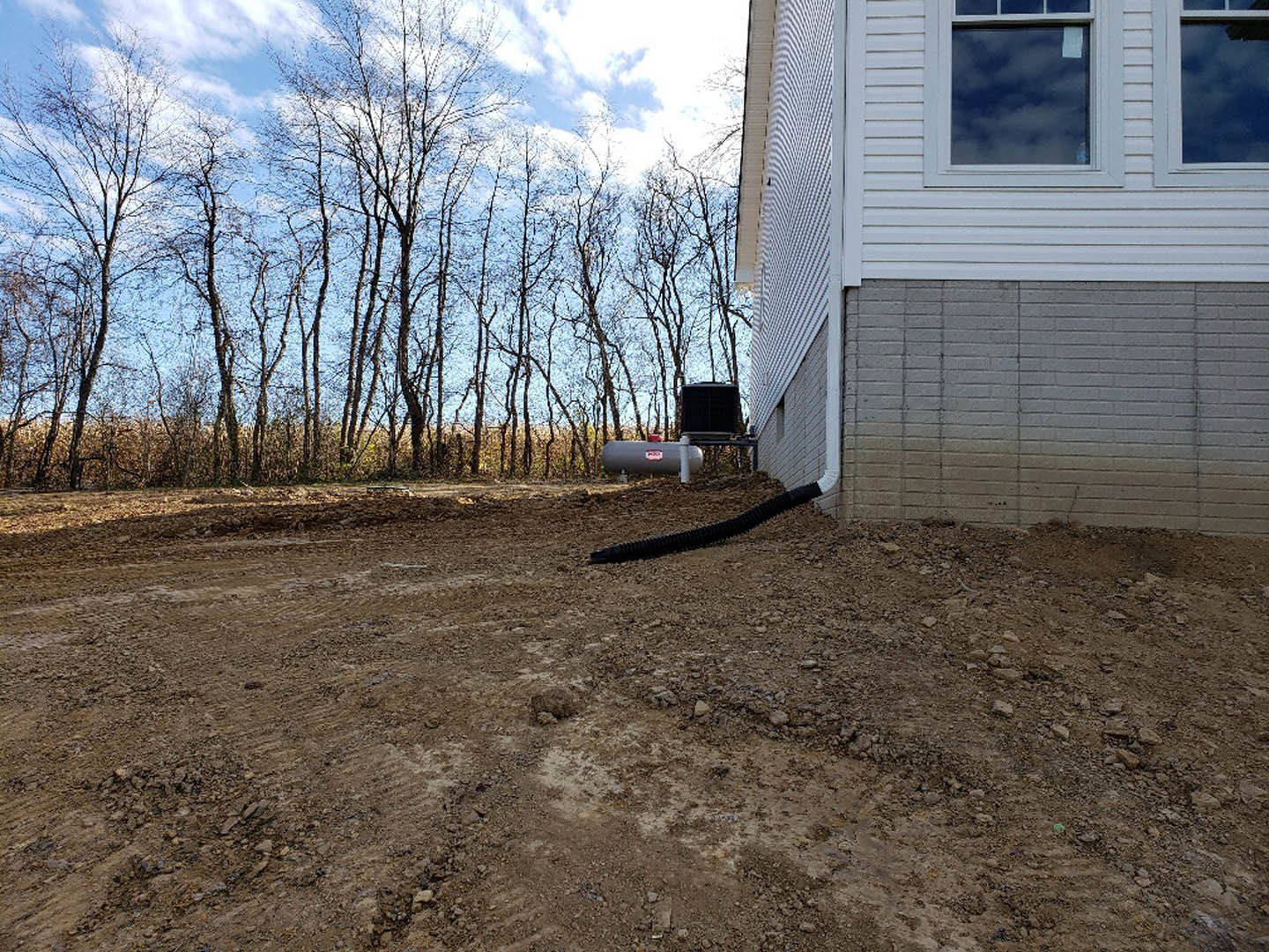 White brick house exterior with black pipe running along dirt ground, leafless trees in background, cloudy sky overhead, window visible on facade.