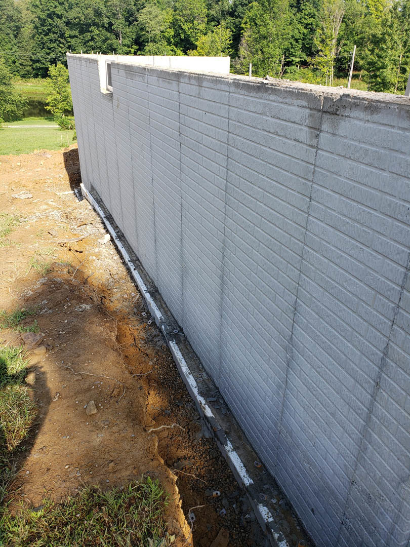 White concrete exterior wall bordered by patchy grass and dirt, circular hole in ground near wall, trees visible in background