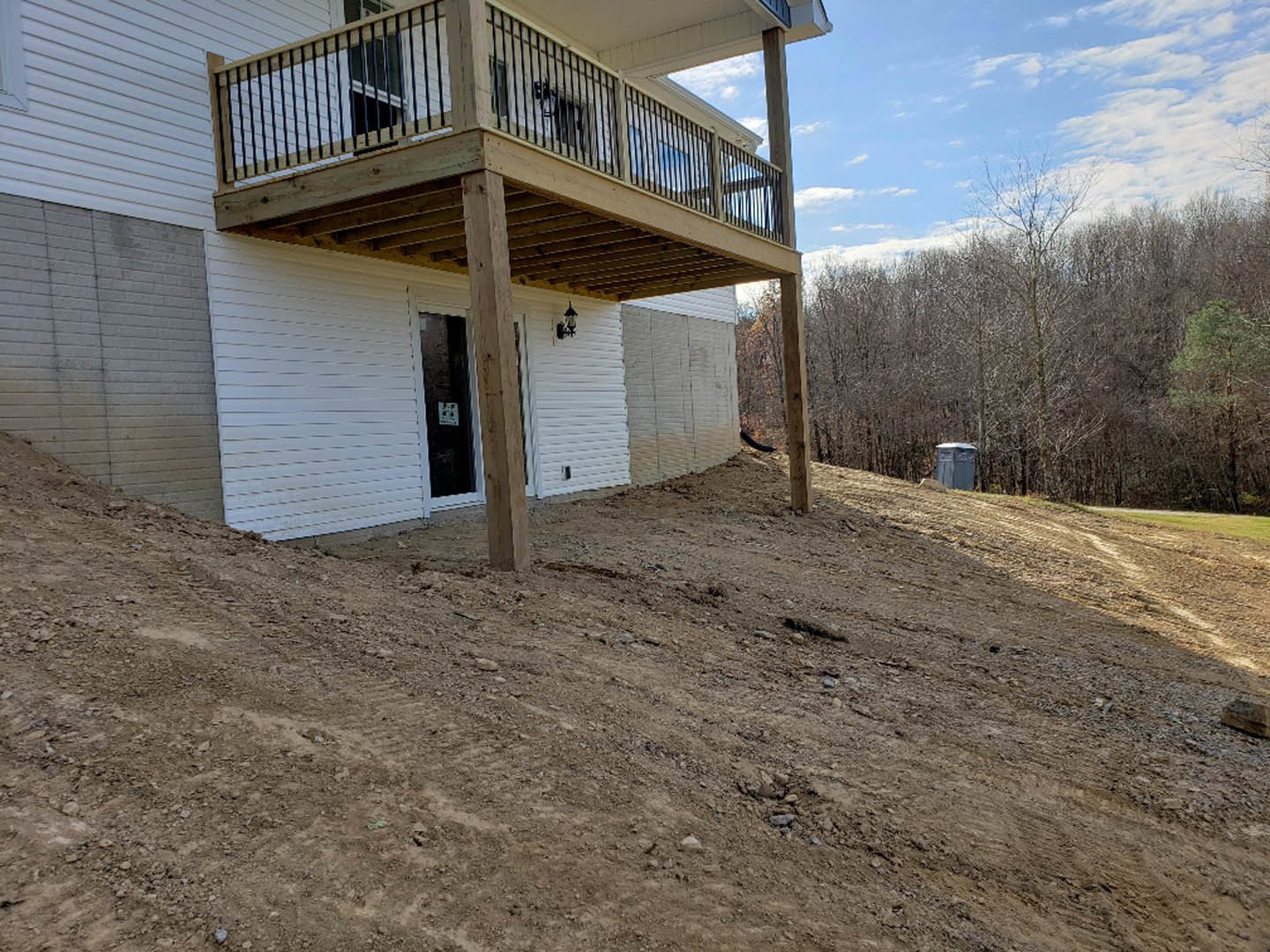 Two-story house with wood balcony and deck, surrounded by trees, dirt hill in foreground, blue portable toilet near wooded area, attached garage with white door, cloudy sky