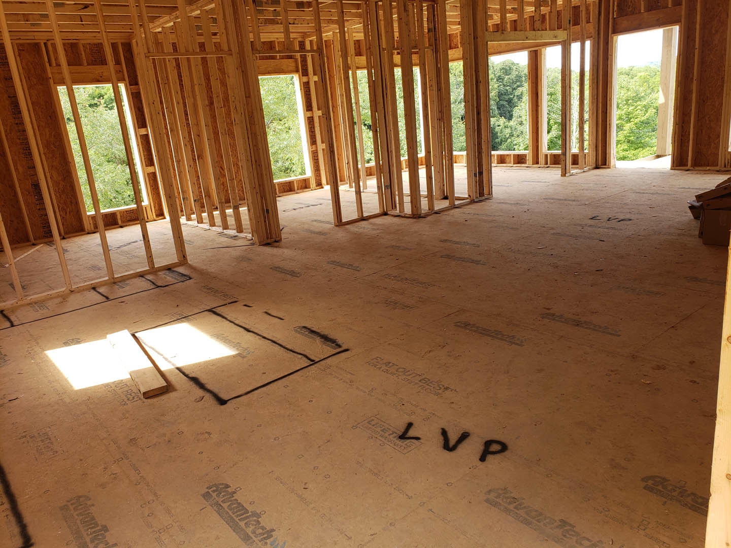 Wood framing and beams forming the floor and walls of a house under construction, sunlight streaming through a window, sandy ground visible, black letter V painted on a surface.
