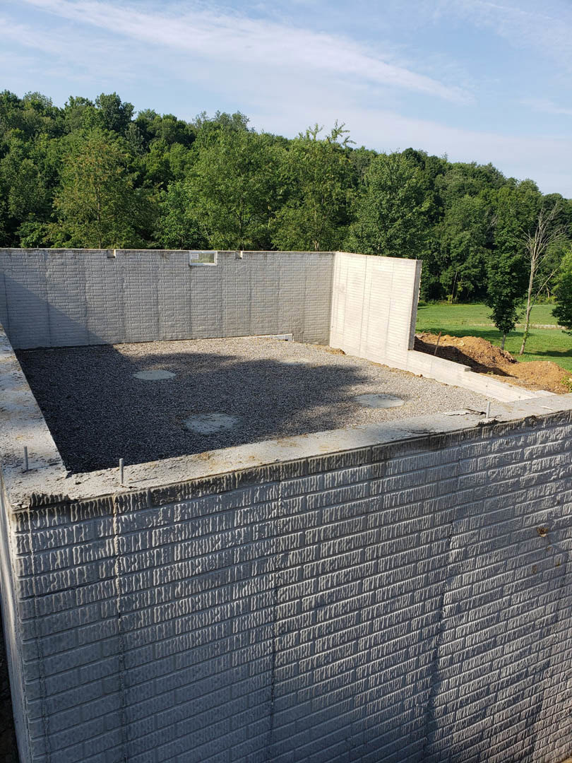 Modern home exterior with grey concrete foundation, large window set in a brick wall, surrounding trees, blue sky, and paved driveway in foreground
