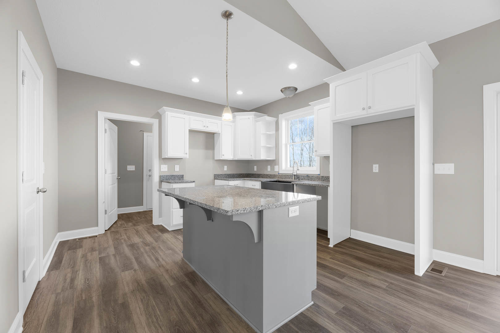 Kitchen with wood flooring, central island featuring granite countertops, white cabinetry, stainless steel sink, bar seating, window overlooking trees, and door with light switch.