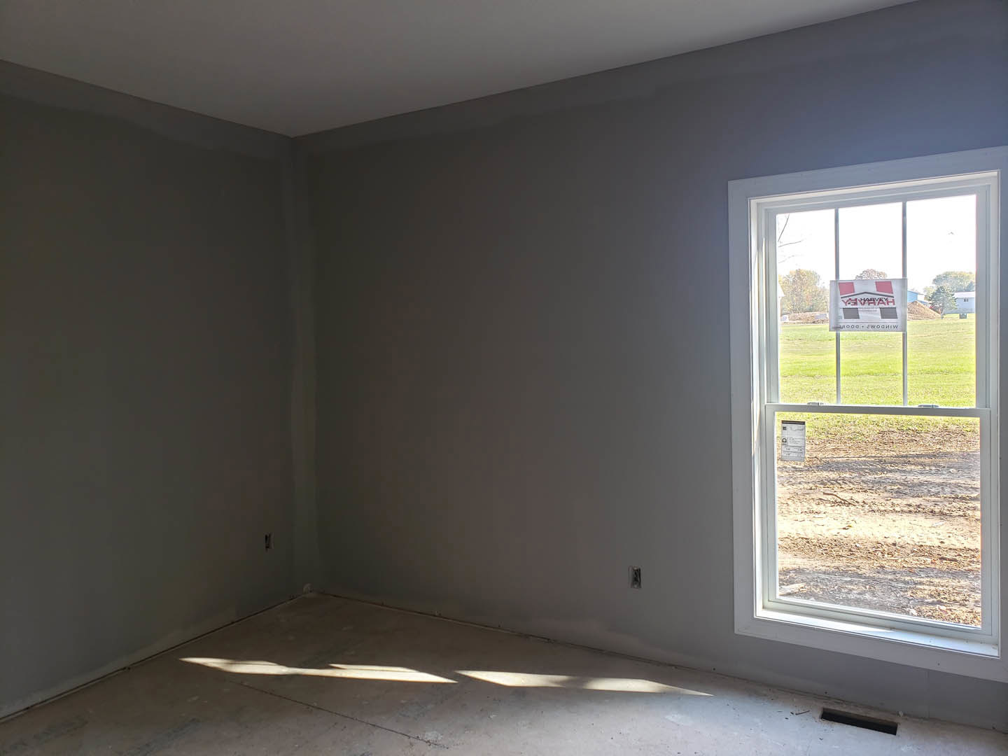 Sunlit room with plaster walls, large window overlooking dirt field and distant tree, sign with text displayed on window, ceiling light fixture illuminating floor.