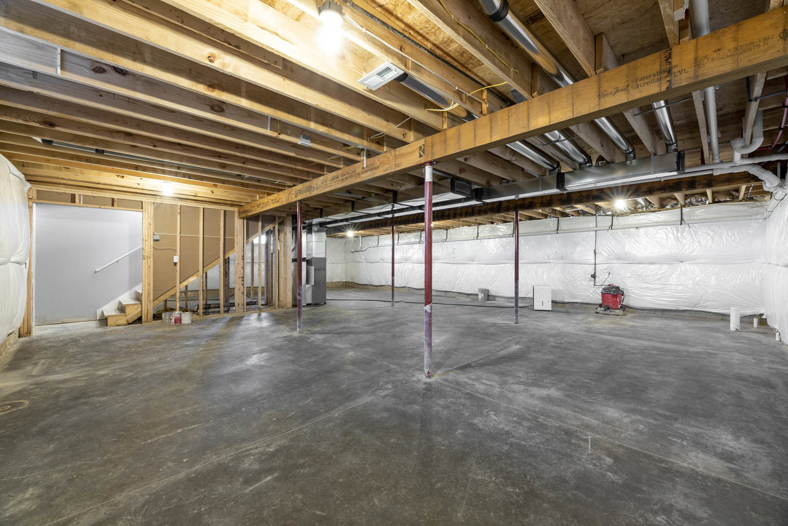 Basement with wooden ceiling, concrete floor, white wall with handrail and vent, red cart with black lid, exposed support pole