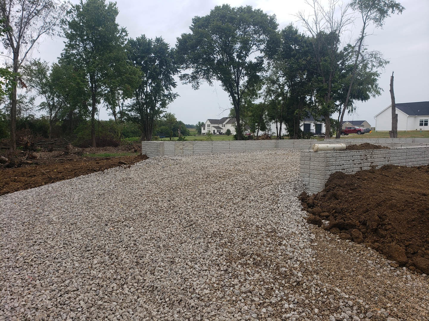 White house with black roof behind gravel road, dirt piles, and stone and brick retaining wall, surrounded by trees and plants.