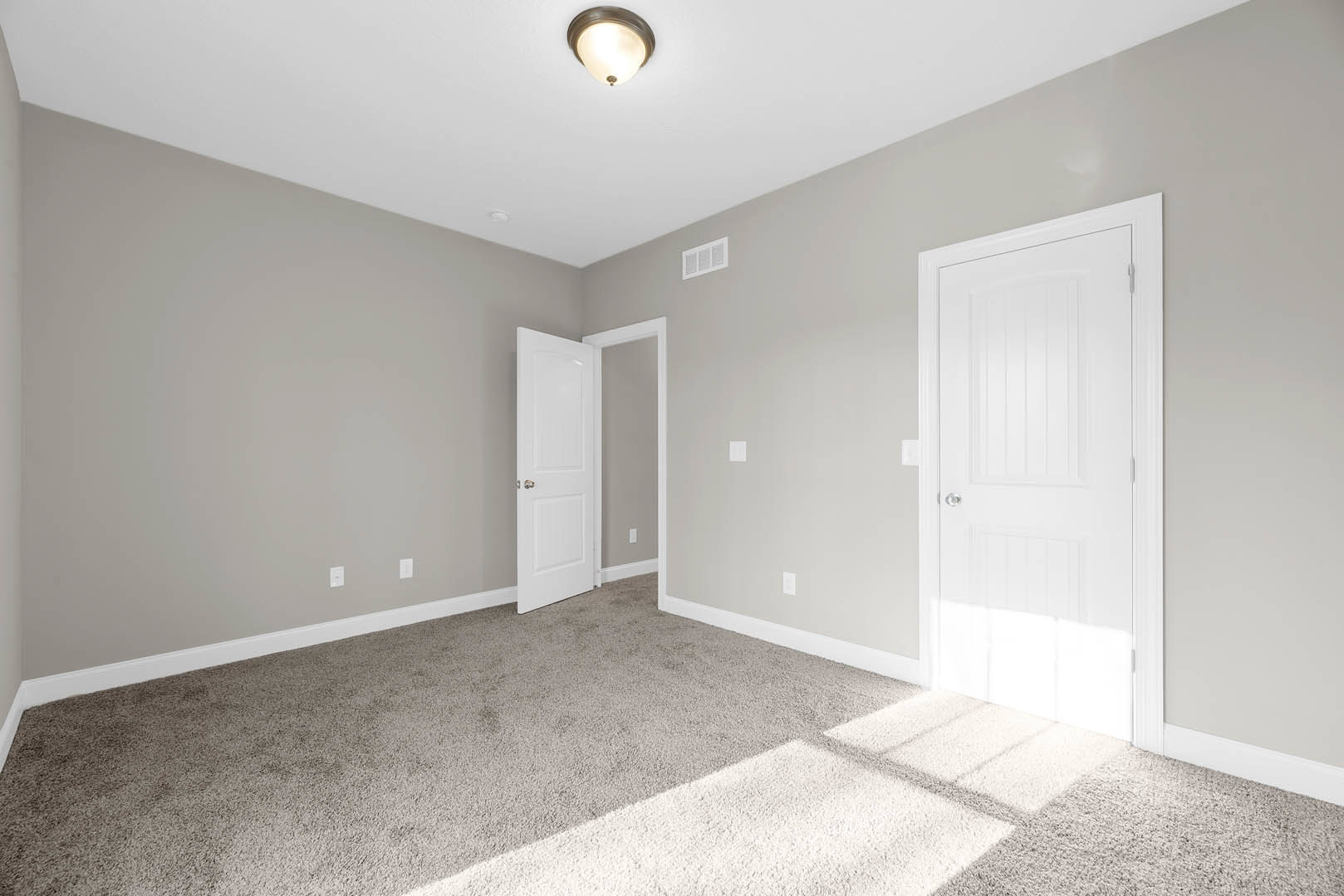 White carpeted floor, white walls with base molding, closed white door featuring a silver knob, ceiling-mounted light fixture, wall vent.