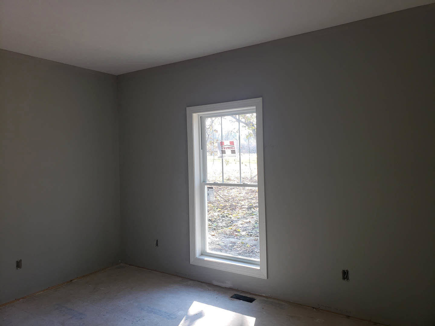 Concrete floor with a corner vent, white plaster walls, large window featuring a sign with red text, partial view of a door casting a shadow.