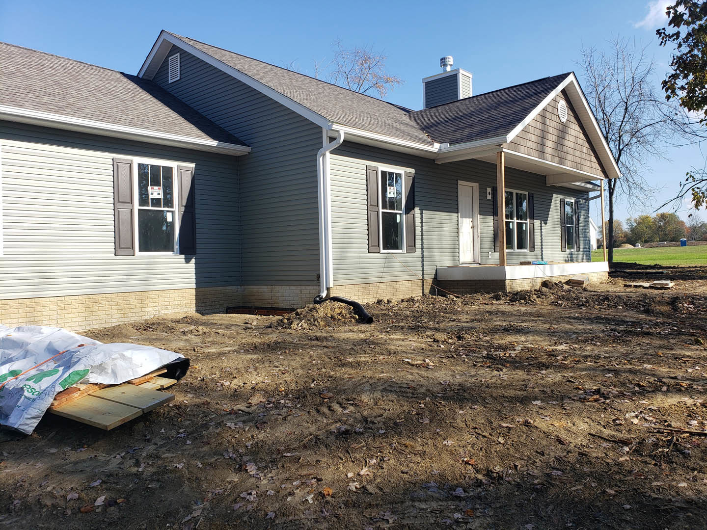 Two-story house with light siding, gutter, and window displaying a sign; dirt yard scattered with debris, exposed pipe, and white tarp covering unfinished wooden surface.